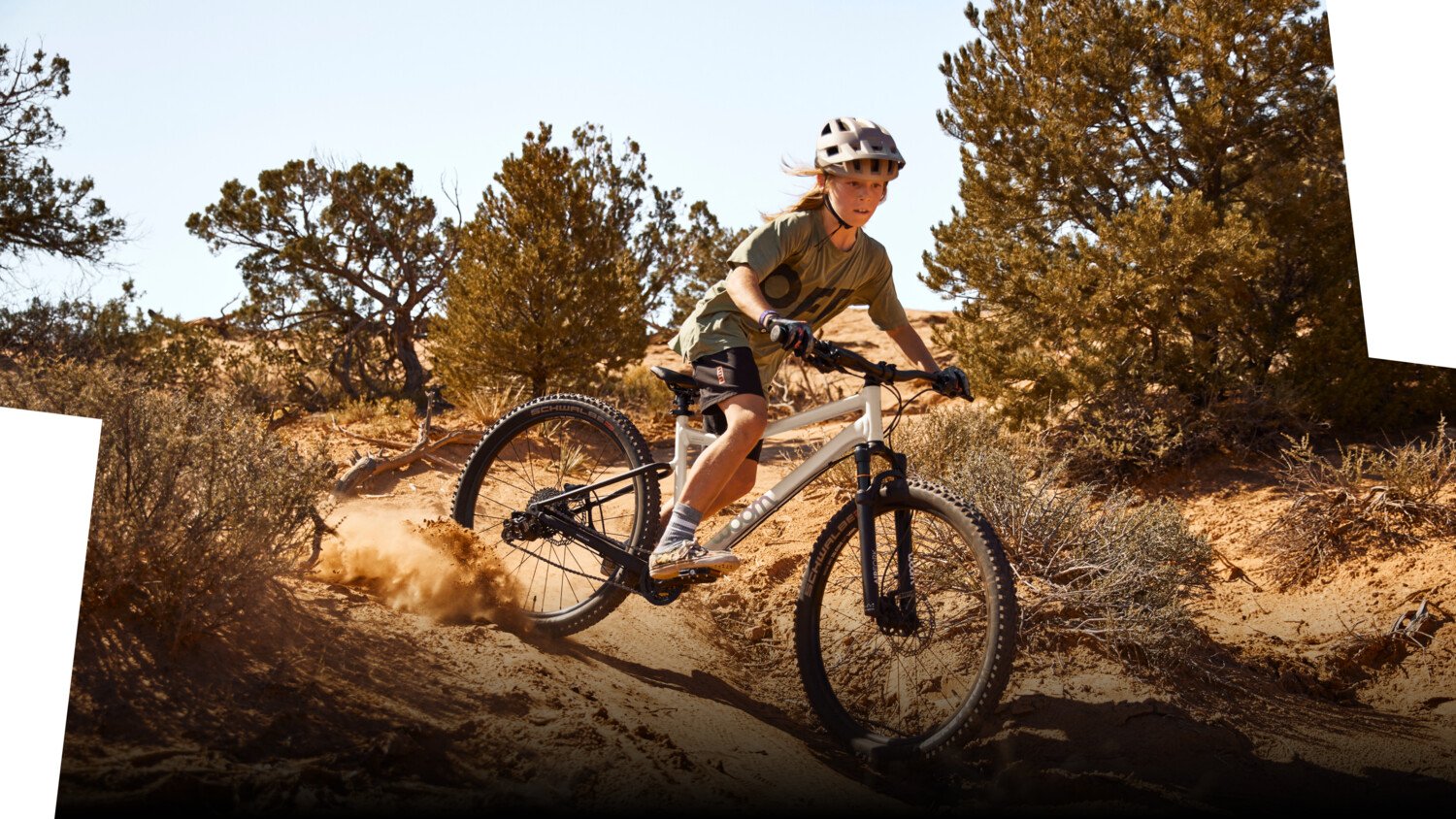 Junge mit Fahrradhelm auf dem Kopf driftet mit einem weißen woom OFF AIR Mountainbike auf einem sandigen Trail.